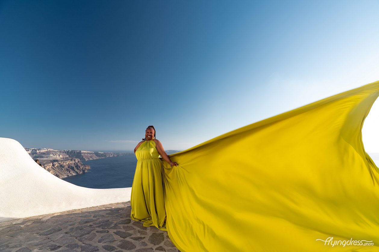 Sunshine meets the sea as a bold yellow flying dress sweeps dramatically across Santorini's iconic cliffside skyline.